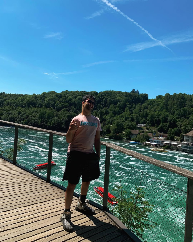 Mert Karataş posing on a wooden pier by a fast-flowing turquoise river, with a forest and a summer sky in the background.